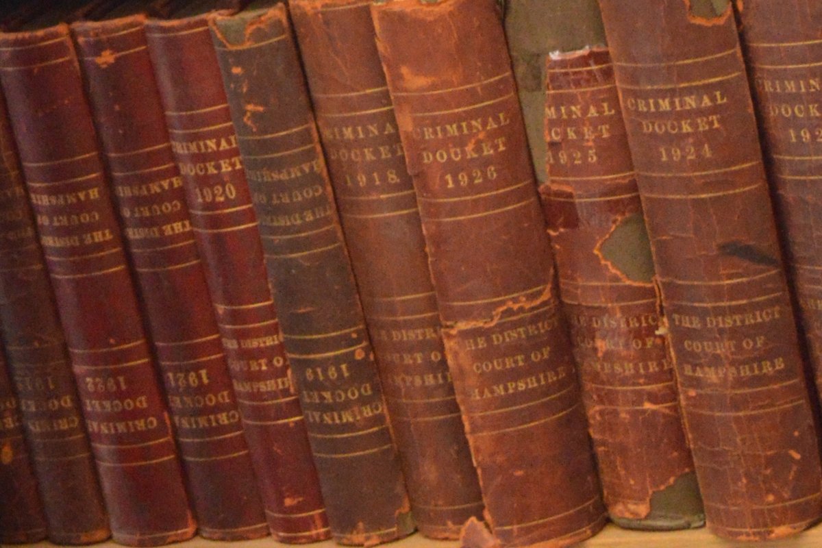 A stack of thick legal law books and statutes documents opened to a page about controlled substances, set against a blurred background of courthouse steps.